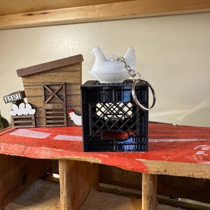 Puede incluir: Un llavero de gallina blanca se encuentra sobre una caja negra, con un llavero plateado. La escena incluye un gallinero de madera en miniatura y un letrero que dice "FRESH". El escenario es una exhibici&oacute;n r&uacute;stica con tem&aacute;tica de granja.