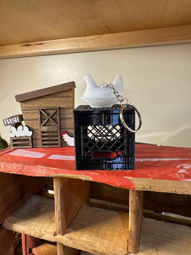 Puede incluir: Un llavero de gallina blanca se encuentra sobre una caja negra, con un llavero plateado. La escena incluye un gallinero de madera en miniatura y un letrero que dice "FRESH". El escenario es una exhibici&oacute;n r&uacute;stica con tem&aacute;tica de granja.