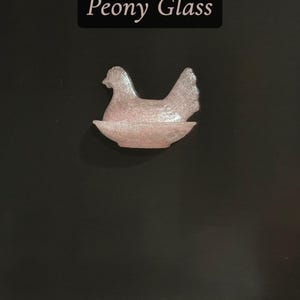 May include: A pink glass hen figurine in a nest shape. The figurine is made of textured glass and sits on a dark background. The text "Peony Glass" is displayed above the figurine.