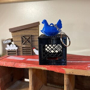 Puede incluir: Un llavero de gallina azul se encuentra sobre una caja de huevos negra llena de huevos blancos. En el fondo, hay un gallinero de madera en miniatura y un letrero que dice "FRESH". El llavero tiene un anillo plateado.