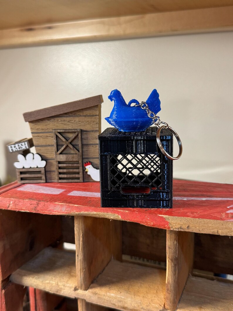 Puede incluir: Un llavero de gallina azul se encuentra sobre una caja de huevos negra llena de huevos blancos. En el fondo, hay un gallinero de madera en miniatura y un letrero que dice "FRESH". El llavero tiene un anillo plateado.