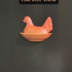 May include: A coral-colored hen figurine in a nest-like bowl. The hen is perched in the center, with a detailed tail and head. The text "Harvest Glow" is displayed in a black rectangle above the hen.