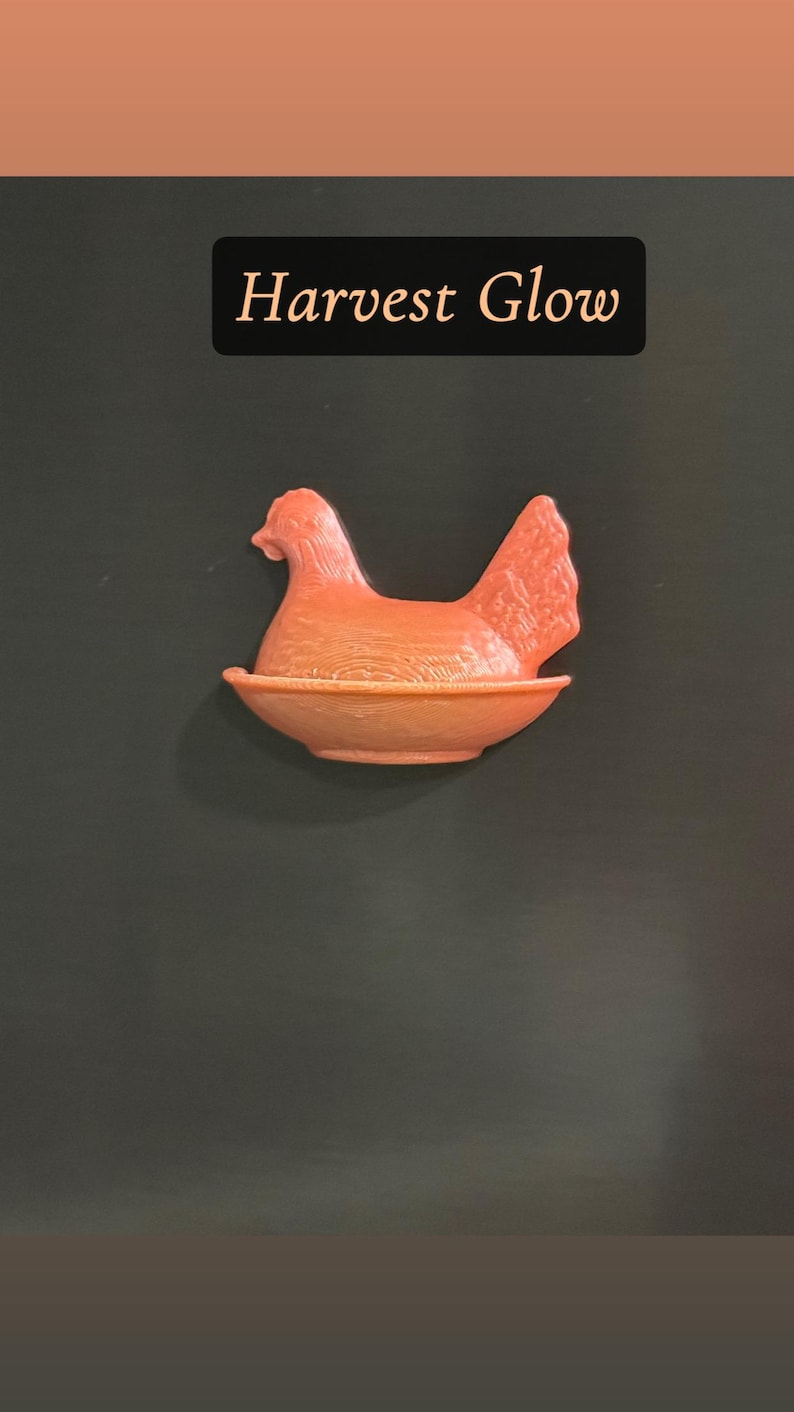 May include: A coral-colored hen figurine in a nest-like bowl. The hen is perched in the center, with a detailed tail and head. The text "Harvest Glow" is displayed in a black rectangle above the hen.