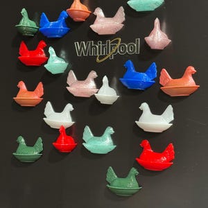 May include: A collection of colorful chicken-shaped magnets in various colors, including red, blue, green, pink, and white. The magnets are arranged on a black surface, with a Whirlpool logo visible. These decorative magnets are likely for the kitchen or home decor.