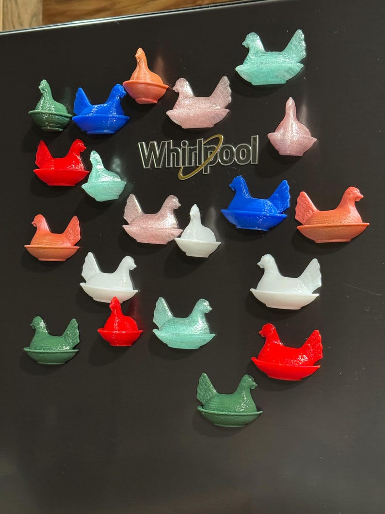 May include: A collection of colorful chicken-shaped magnets in various colors, including red, blue, green, pink, and white. The magnets are arranged on a black surface, with a Whirlpool logo visible. These decorative magnets are likely for the kitchen or home decor.