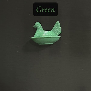 May include: A green hen figurine in a nest-like bowl. The word "Green" is displayed in a black rectangle above the figurine. The background is a dark gray color.
