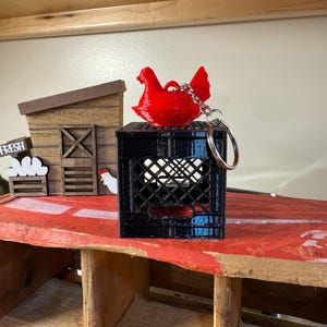Puede incluir: Un llavero de gallina roja sobre una caja negra. El llavero est&aacute; unido a un anillo plateado. En el fondo hay una estructura de madera con la palabra "FRESH" y una peque&ntilde;a ilustraci&oacute;n de gallina.