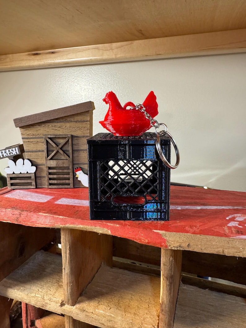 Puede incluir: Un llavero de gallina roja sobre una caja negra. El llavero est&aacute; unido a un anillo plateado. En el fondo hay una estructura de madera con la palabra "FRESH" y una peque&ntilde;a ilustraci&oacute;n de gallina.