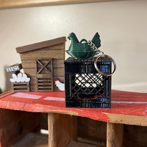 Puede incluir: Un llavero de pollo verde se encuentra sobre una mini caja negra. Un peque&ntilde;o gallinero de madera y un letrero que dice "FRESH" est&aacute;n en el fondo. El llavero tiene un anillo plateado.
