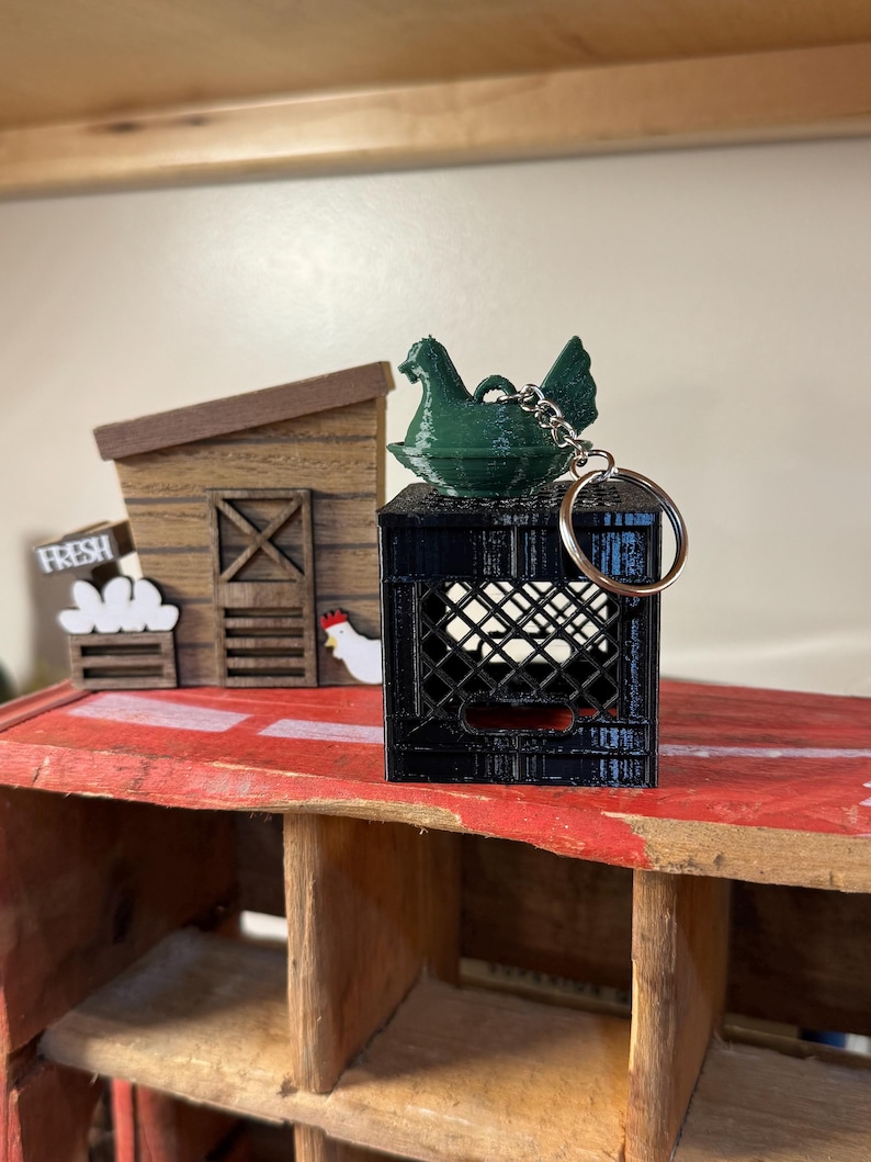 Puede incluir: Un llavero de pollo verde se encuentra sobre una mini caja negra. Un peque&ntilde;o gallinero de madera y un letrero que dice "FRESH" est&aacute;n en el fondo. El llavero tiene un anillo plateado.