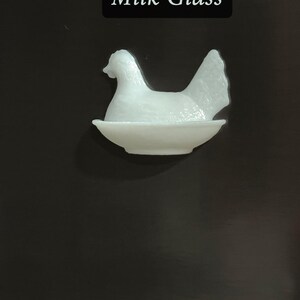 May include: A white milk glass hen figurine sitting in a shallow dish. The hen is detailed with a textured surface and is set against a dark background. The text "Milk Glass" is displayed in a black rectangle.