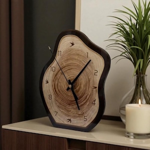 Può includere: Un orologio in stile legno con cornice marrone scuro e quadrante marrone chiaro. Il quadrante presenta un design ad anelli di albero con lancette e numeri neri. L'orologio è su una superficie bianca.