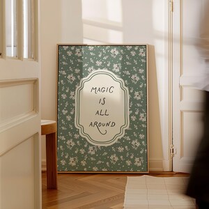 K&ouml;nnte beinhalten: Ein gerahmter Wandkunstdruck mit einem gr&uuml;nen floralen Hintergrund. Das Kunstwerk zeigt ein wei&szlig;es, ornatenf&ouml;rmiges zentrales Design mit den Worten "MAGIC IS ALL AROUND" in schwarzer Schrift. Der Rahmen ist hellbraun.