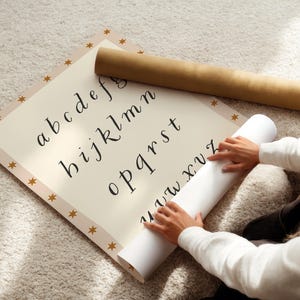 May include: A cream-colored alphabet chart with a gold star border and a rolled-up paper. The chart displays lowercase letters in a handwritten style. A brown paper tube rests on top of the chart.