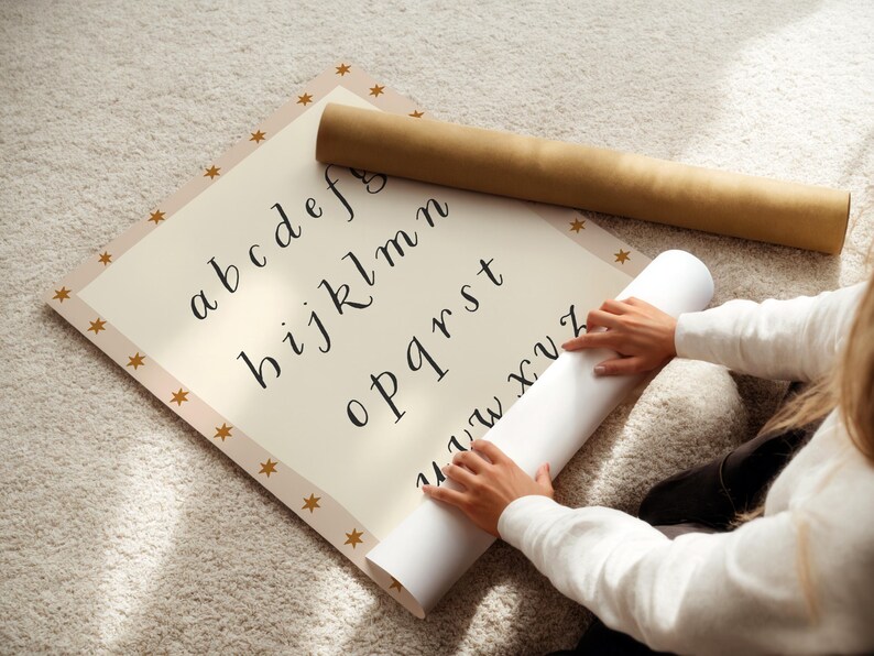 May include: A cream-colored alphabet chart with a gold star border and a rolled-up paper. The chart displays lowercase letters in a handwritten style. A brown paper tube rests on top of the chart.