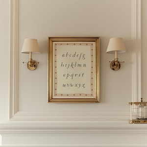May include: A framed print of the alphabet in a gold frame, hanging on a wall with two sconces. The print has a cream background with the letters a through z in a handwritten style. The frame is rectangular with a gold finish.