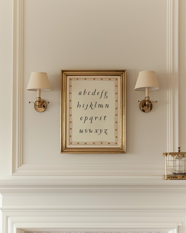 May include: A framed print of the alphabet in a gold frame, hanging on a wall with two sconces. The print has a cream background with the letters a through z in a handwritten style. The frame is rectangular with a gold finish.