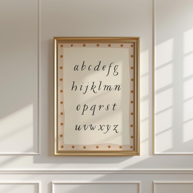 May include: A framed print featuring the lowercase alphabet in a delicate, handwritten font. The print is set against a cream-colored background and framed in gold with star accents. The artwork is displayed on a white wall.
