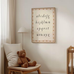 May include: A framed alphabet print in a nursery setting. The print features the lowercase alphabet in a cursive font, with a star-patterned border. A brown teddy bear sits on a wooden chair with a cream-colored pillow. A floor lamp and crib are also visible.