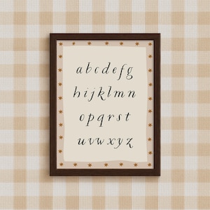 May include: A framed print featuring the lowercase alphabet in a stylish, handwritten font. The print is set against a cream-colored background with a border of small gold stars. The frame is a dark brown color, and the background is a beige and white checkered pattern.