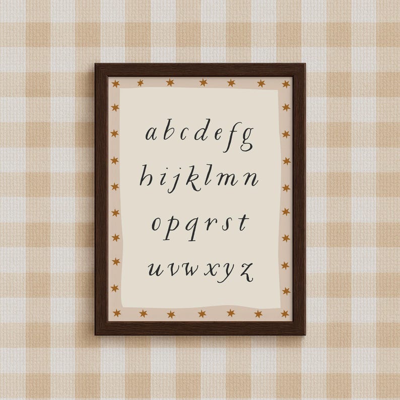 May include: A framed print featuring the lowercase alphabet in a stylish, handwritten font. The print is set against a cream-colored background with a border of small gold stars. The frame is a dark brown color, and the background is a beige and white checkered pattern.