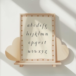 May include: A framed alphabet print in a light wood frame with a star-patterned border. The print features the lowercase alphabet in a delicate, handwritten font on a cream-colored background. The frame is displayed on a cloud-shaped shelf.