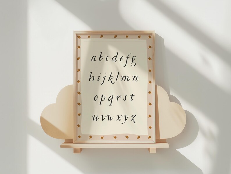 May include: A framed alphabet print in a light wood frame with a star-patterned border. The print features the lowercase alphabet in a delicate, handwritten font on a cream-colored background. The frame is displayed on a cloud-shaped shelf.