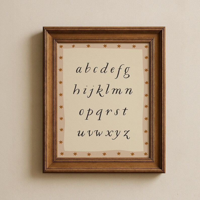 May include: A framed print featuring the lowercase alphabet in a cursive font. The letters are black on a cream-colored background. The frame is a dark brown wood, and the print is bordered with small gold stars.