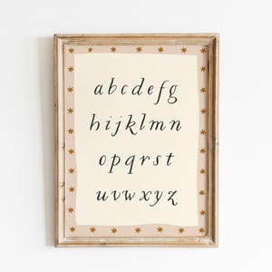 May include: A framed print featuring the lowercase alphabet in a handwritten style. The letters are in black on a cream-colored background. The frame is light brown with a border of small gold stars.