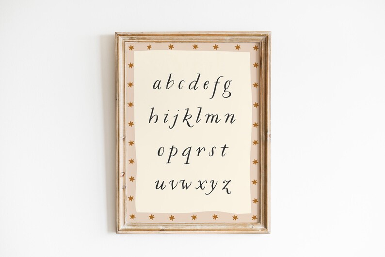 May include: A framed print featuring the lowercase alphabet in a handwritten style. The letters are in black on a cream-colored background. The frame is light brown with a border of small gold stars.