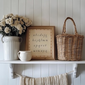 May include: A decorative shelf displays a framed alphabet print, a woven basket, a ceramic vase with flowers, and a small mug. The alphabet print is in a wooden frame with star accents. The vase and mug are cream-colored.