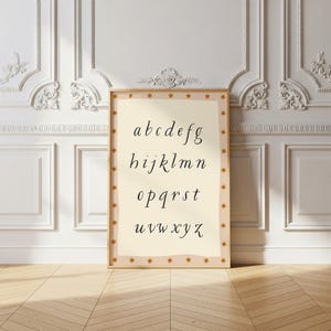 May include: A framed print featuring the alphabet in a cursive font. The letters are in black on a cream-colored background. The frame is a light brown with small gold stars around the border. The print is leaning against a white wall with decorative molding.