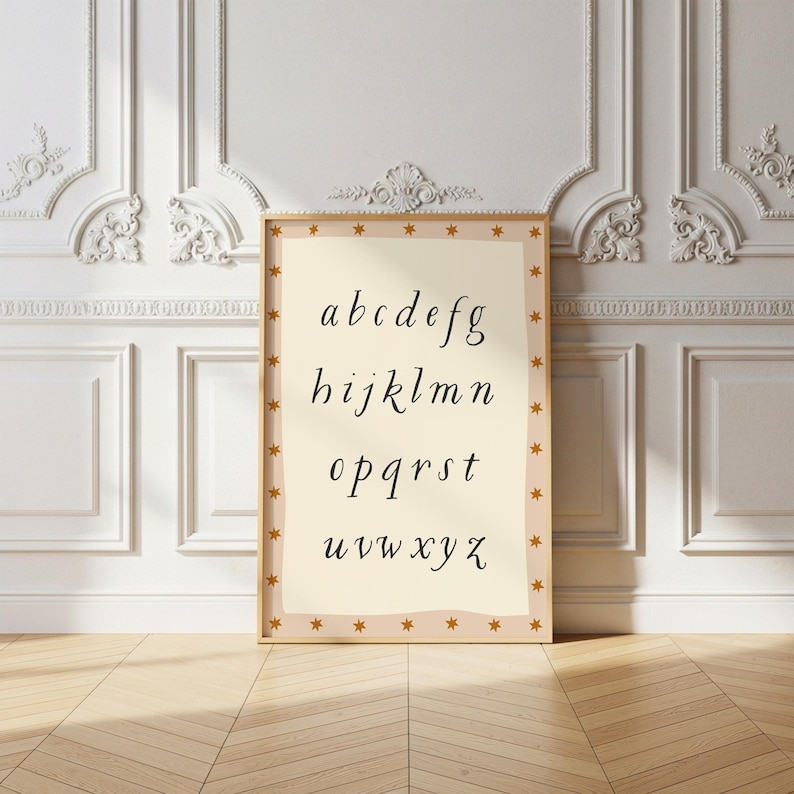 May include: A framed print featuring the alphabet in a cursive font. The letters are in black on a cream-colored background. The frame is a light brown with small gold stars around the border. The print is leaning against a white wall with decorative molding.