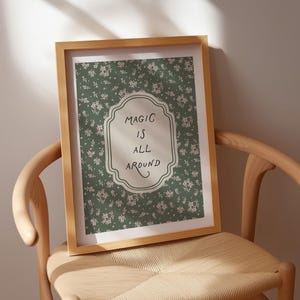 K&ouml;nnte beinhalten: Ein gerahmter Druck mit den Worten "MAGIC IS ALL AROUND" in einem dekorativen Rahmen. Der Druck hat einen gr&uuml;nen floralen Hintergrund. Der Rahmen besteht aus hellbraunem Holz, und der Druck wird auf einem Holzstuhl pr&auml;sentiert.