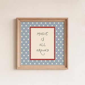 May include: A framed print featuring the phrase "MAGIC IS ALL AROUND" in black script. The background is pale blue with white polka dots. The text is on a cream rectangle with a red scalloped border, set in a light brown wooden frame.