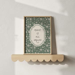 K&ouml;nnte beinhalten: Ein gerahmter Druck mit den Worten "MAGIC IS ALL AROUND" in einem dekorativen Rahmen, vor einem gr&uuml;nen floralen Hintergrund. Der Druck wird auf einem wellenf&ouml;rmigen Holzregal pr&auml;sentiert, was eine charmante, skurrile &Auml;sthetik erzeugt.