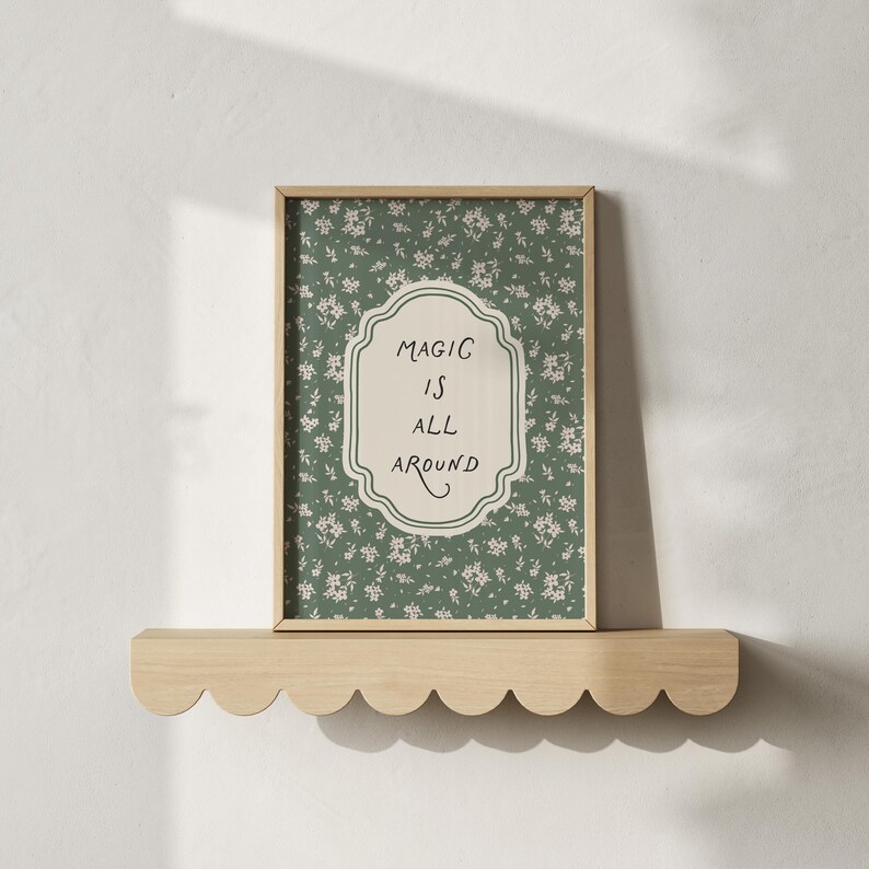 K&ouml;nnte beinhalten: Ein gerahmter Druck mit den Worten "MAGIC IS ALL AROUND" in einem dekorativen Rahmen, vor einem gr&uuml;nen floralen Hintergrund. Der Druck wird auf einem wellenf&ouml;rmigen Holzregal pr&auml;sentiert, was eine charmante, skurrile &Auml;sthetik erzeugt.