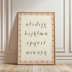 May include: A framed print featuring the lowercase alphabet in a stylish, handwritten font. The letters are black on a cream-colored background, framed by a border of small gold stars. The frame is a warm, natural wood tone.