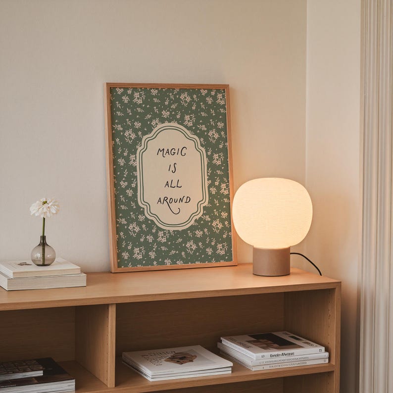 K&ouml;nnte beinhalten: Ein Holzregal zeigt einen gerahmten Blumendruck mit den Worten "MAGIC IS ALL AROUND", eine kugelf&ouml;rmige Tischlampe, eine kleine Vase mit einer Blume und gestapelte B&uuml;cher. Die Lampe hat einen beigefarbenen Sockel und einen wei&szlig;en, abgerundeten Schirm.