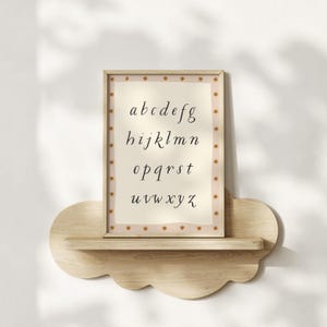 May include: A framed alphabet print with a beige background and a star-patterned border. The letters a through z are displayed in a cursive font. The frame is displayed on a cloud-shaped wooden shelf.