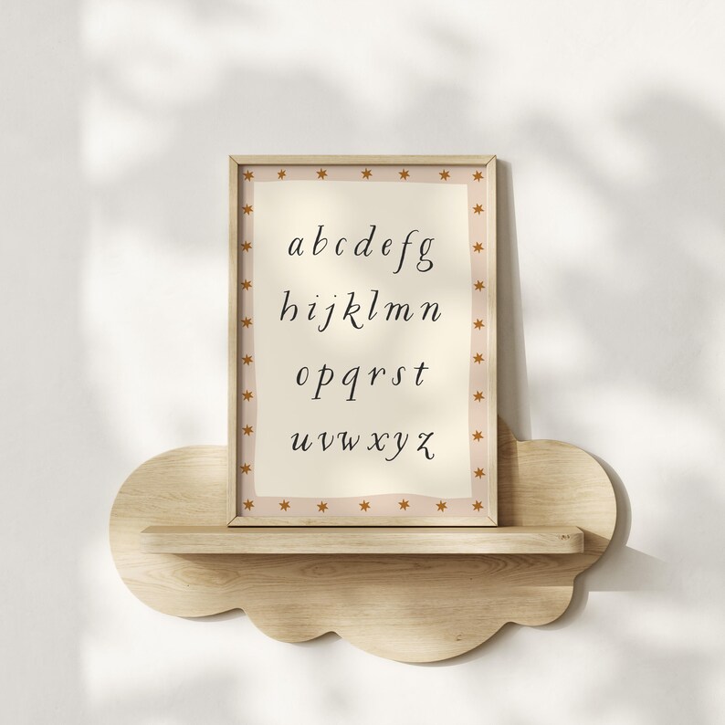 May include: A framed alphabet print with a beige background and a star-patterned border. The letters a through z are displayed in a cursive font. The frame is displayed on a cloud-shaped wooden shelf.