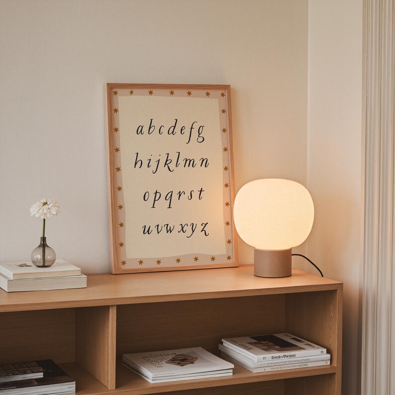 May include: A wooden shelf displays a framed alphabet print, a glowing table lamp, and a small vase with a flower. The alphabet print has a light background with the letters a-z in a handwritten style. The lamp has a round, white shade.