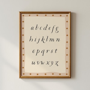 May include: A framed print featuring the lowercase alphabet in a cursive font. The letters are black on a cream-colored background. The frame is gold, with a border of small gold stars. The print is a decorative piece.