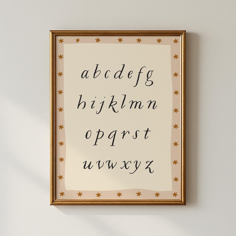 May include: A framed print featuring the lowercase alphabet in a cursive font. The letters are black on a cream-colored background. The frame is gold, with a border of small gold stars. The print is a decorative piece.