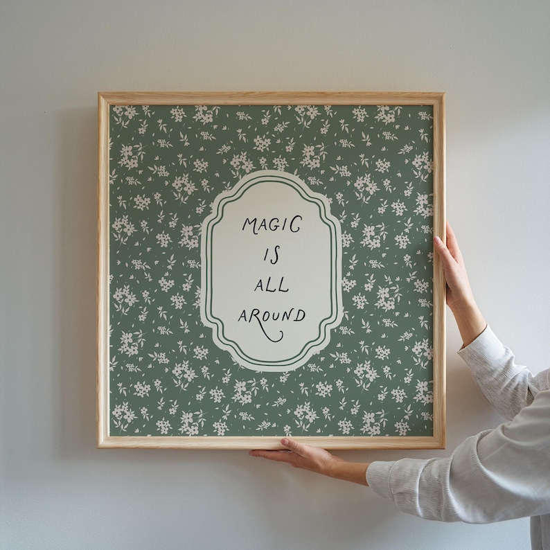 K&ouml;nnte beinhalten: Ein gerahmter Druck mit einem gr&uuml;nen floralen Hintergrundmuster und den Worten "MAGIC IS ALL AROUND" in einem wei&szlig;en, dekorativen Rahmen. Der Rahmen besteht aus hellem Holz.