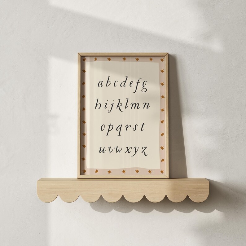May include: A framed alphabet print with a beige background and a border of gold stars. The letters a through z are displayed in a black, elegant font. The frame is displayed on a light wood shelf with a scalloped edge.