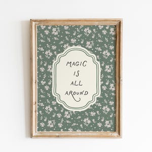 K&ouml;nnte beinhalten: Gerahmtes Wandbild mit floralem Muster in Gr&uuml;n und Wei&szlig;. Das Kunstwerk zeigt ein cremefarbenes, verziertes Etikett mit den Worten "MAGIC IS ALL AROUND" in schwarzer Schrift. Der Rahmen ist hellbraun.