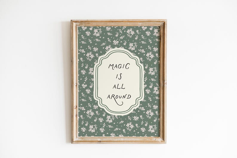 K&ouml;nnte beinhalten: Gerahmtes Wandbild mit floralem Muster in Gr&uuml;n und Wei&szlig;. Das Kunstwerk zeigt ein cremefarbenes, verziertes Etikett mit den Worten "MAGIC IS ALL AROUND" in schwarzer Schrift. Der Rahmen ist hellbraun.