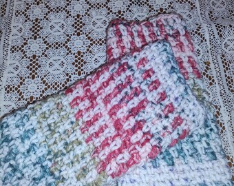 May include: Close-up of three handmade crochet items in shades of white, pink, blue, and green. The items appear to be small, rectangular, and possibly intended for use as wrist warmers or similar accessories. The crochet stitch creates a textured, striped pattern.