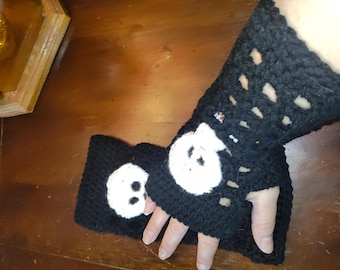 May include: Black crochet fingerless gloves with a white skull design. The gloves are handmade and feature a textured pattern with openwork details. The skull is centered on the back of the hand.